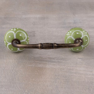 May include: A green ceramic drawer pull with a white floral design. The pull has a brass bar connecting the two knobs.