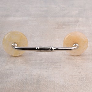 May include: A cabinet handle featuring a polished silver-tone metal bar connected to two circular, translucent, pale yellow stone knobs. The metal bar has a smooth, curved design with a central decorative detail, connecting the two stone ends.