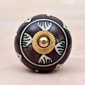 May include: A round ceramic cabinet knob featuring a dark brown base with a white hand-painted fan-like pattern. The centre of the knob is adorned with a circular gold-toned metal hardware piece that has a ribbed edge and a smooth, raised dome.