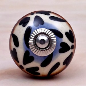 May include: A round ceramic cabinet knob featuring a cream-coloured base with a hand-painted pattern of black heart-shaped leaves and blue accents. The centre of the knob has a silver-toned metal hardware fitting with a ridged circular edge.