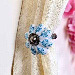 May include: A white ceramic curtain tieback featuring a scalloped, flower-like shape with a blue leaf pattern. The center of the tieback has a dark, metallic, circular accent with a ridged edge, securing a piece of light-colored, textured fabric.