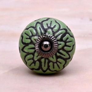 May include: A round ceramic cabinet knob featuring a light green base colour with a dark grey floral pattern. The centre of the knob has a circular metallic silver-toned hardware piece with a ridged edge and a small central screw hole.