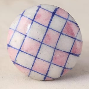 May include: A round ceramic knob with a pink and blue checkered pattern. The knob is white with a glossy finish.