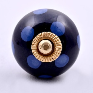 May include: A spherical cabinet knob with a glossy black finish and a pattern of light blue polka dots. The center of the knob features a circular gold-toned metal hardware component with a ridged edge and a smooth, raised central post.