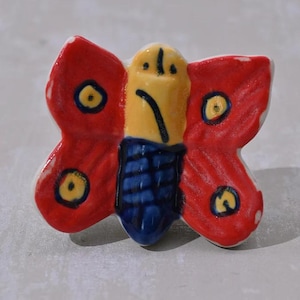 Könnte beinhalten: Eine Keramik-Schmetterlingsfigur mit roten Flügeln, einem gelben Körper und blauen Details. Der Schmetterling hat ein einfaches, handgemaltes Design mit gelben Kreisen auf den Flügeln.