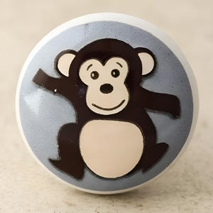 May include: A ceramic knob with a blue background and a cartoon monkey design. The monkey is brown with a light brown belly and a white face.
