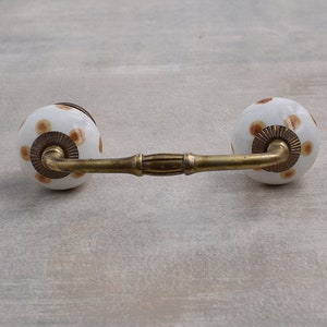 May include: White ceramic drawer pull with brown polka dots and a brass bar connecting the two knobs.