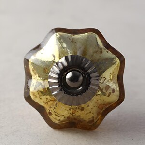 Op de afbeelding: Een decoratieve kastknop met een stervormig ontwerp. De knop heeft een goudkleurig, getextureerd glazen midden met een donkerbruine rand. Het midden is omgeven door een donkere metalen basis en een kleine, donkere metalen bal.
