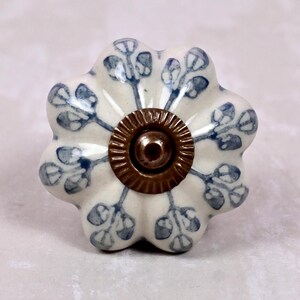 May include: A flower-shaped ceramic cabinet knob with a cream-coloured base and a blue painted leaf pattern. The centre of the knob features a circular, ribbed, antique bronze-coloured metal hardware piece with a small protruding screw head.