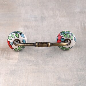 May include: A ceramic drawer pull with a floral design in shades of red, green, and blue. The pull has a brass bar connecting the two ceramic knobs.