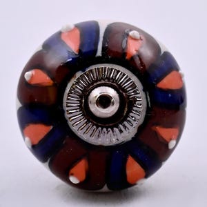 May include: A round ceramic cabinet knob featuring a repeating pattern of dark brown and deep blue petals with bright orange accents and small white dots. The centre of the knob has a circular silver-toned metal hardware piece with ridges.