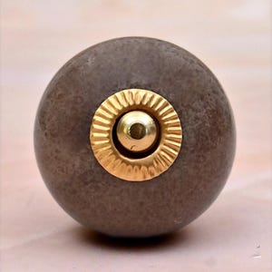 Op de afbeelding: Een bolvormige meubelknop met een getextureerde, matte bruine afwerking. Het midden van de knop is voorzien van een ronde, gepolijste goudkleurige metalen plaat met een geribbelde, zonvormige rand rondom een kleine, gladde koepel.