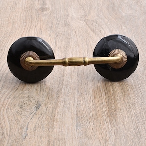 May include: Black ceramic drawer pulls with a gold metal connecting bar. The pulls have a decorative design around the edge.