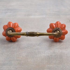 May include: Orange ceramic drawer pull with a brass curved bar connecting two knobs. The knobs are shaped like a flower with a raised center.