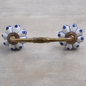 Op de afbeelding: Een witte keramische ladegreep met blauwe stippen en een gouden metalen balk die de twee zijden verbindt. De greep heeft de vorm van een bloem met 8 bloemblaadjes.