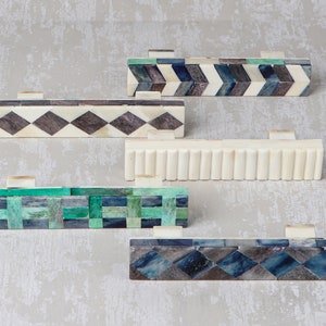 Puede incluir: Cinco tiradores de cajones decorativos hechos de hueso e incrustados con patrones geométricos coloridos. Los tiradores presentan una variedad de diseños, incluyendo patrones de chevron, diamante y cuadros. Los tiradores son todos en tonos de azul, verde y negro.