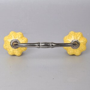 May include: A pair of yellow ceramic drawer pulls with a silver metal connecting bar. The pulls are shaped like a flower with a textured surface.