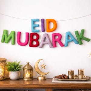 May include: A colorful felt banner spelling "EID MUBARAK" hangs against a white wall. The letters are in various colors: green, pink, red, purple, blue, and orange. Decorative items, including a gold crescent moon and star, are on a wooden surface below.