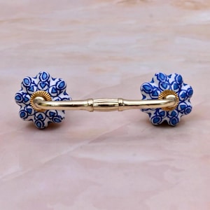 May include: A drawer pull featuring a polished gold-tone metal handle connected to two flower-shaped ceramic knobs. The knobs are white with a detailed blue floral pattern and gold-tone circular accents where the handle meets the ceramic base.
