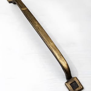 Puede incluir: Un tirador de armario de metal dorado con un diseño cuadrado en cada extremo. El tirador mide aproximadamente 15 cm de largo.