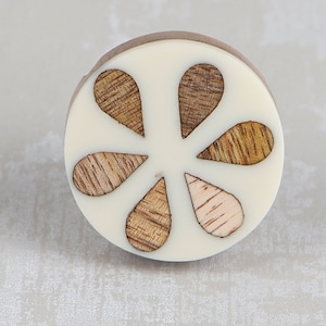 May include: A white round knob with a flower design made of wood. The flower has six petals, each with a different wood grain pattern.