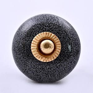 May include: A round cabinet knob with a dark gray and black speckled finish. The center features a circular gold-toned metal accent with a fluted, ridged edge surrounding a smooth, raised dome in the middle of the hardware piece.