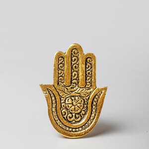 May include: A gold-colored Hamsa hand-shaped decorative item. The hand features intricate floral and scrollwork designs. The item is set against a plain white background.
