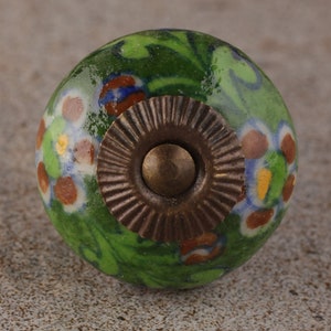 Könnte beinhalten: Ein dekorativer Keramik-Schrankknopf mit floralem Design. Der Knopf ist hauptsächlich grün mit farbenfrohen floralen Akzenten in Blau-, Braun- und Weißtönen. Er hat eine bronzefarbene Metallbasis.