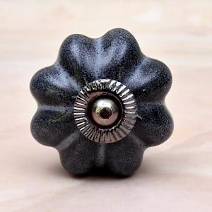 May include: A dark grey ceramic cabinet knob with a scalloped, flower-like shape and a speckled finish. The centre features a circular, silver-toned metal plate with a ridged edge and a smooth, raised dome in the middle of the hardware piece.