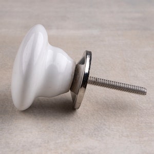 May include: A white ceramic cabinet knob with a smooth, rounded mushroom-shaped head. The knob features a polished silver-coloured metal base and a threaded metal bolt extending from the back for installation on furniture or cabinetry doors.