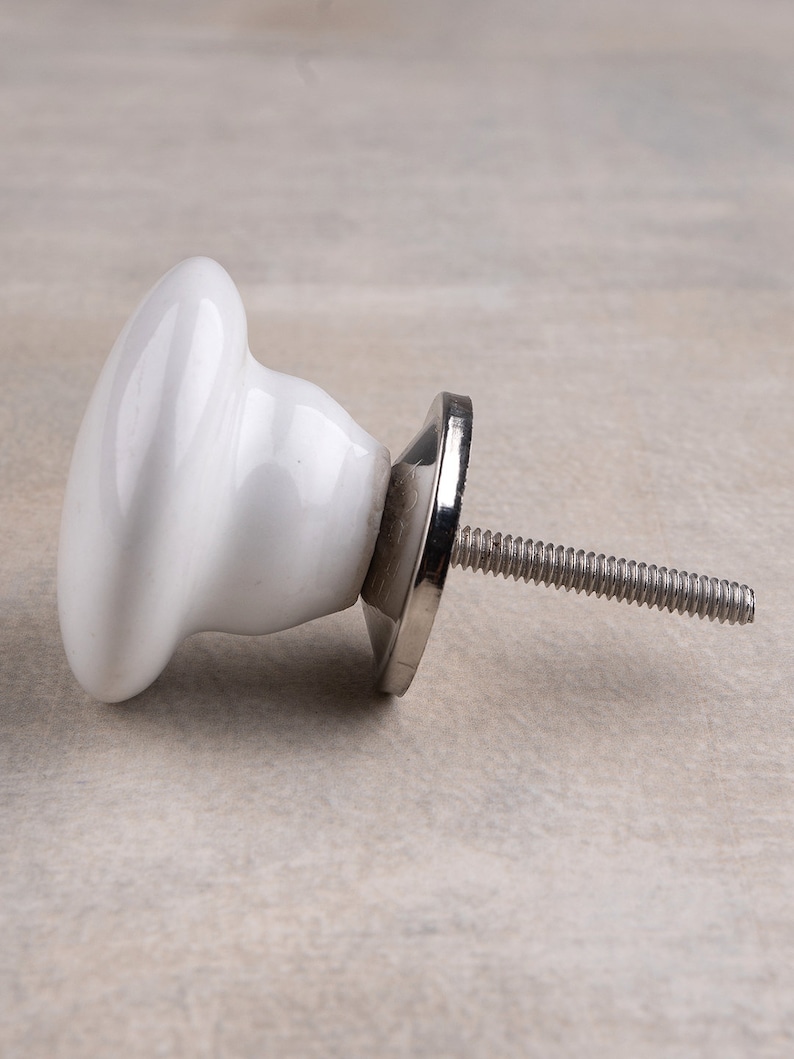 May include: A white ceramic cabinet knob with a smooth, rounded mushroom-shaped head. The knob features a polished silver-coloured metal base and a threaded metal bolt extending from the back for installation on furniture or cabinetry doors.