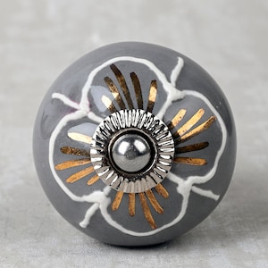 Puede incluir: Pomo de cerámica gris con un diseño de flor blanca y detalles dorados. El pomo tiene una base de metal plateado.