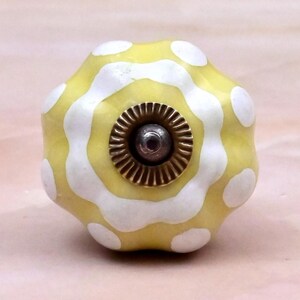 Op de afbeelding: Een ronde, geschulpte keramische meubelknop met een geel en wit golvend streeppatroon. Het midden van de knop heeft een cirkelvormig, bronskleurig metalen onderdeel met een geribbelde rand en een kleine, donkere centrale schroefkop.