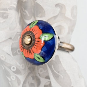Puede incluir: Un sujetalibros de cortina de cerámica azul con una flor naranja pintada a mano y hojas verdes. El sujetalibros tiene un anillo de metal plateado.
