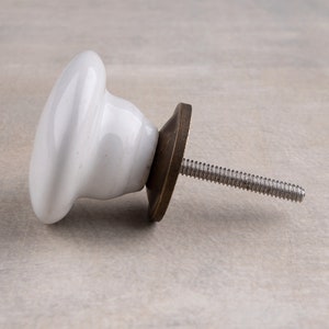 May include: A white ceramic cabinet knob with a smooth, rounded surface and a flared base. The knob features a dark, metallic bronze-toned circular backplate and a long, threaded metal screw extending from the centre for installation purposes.