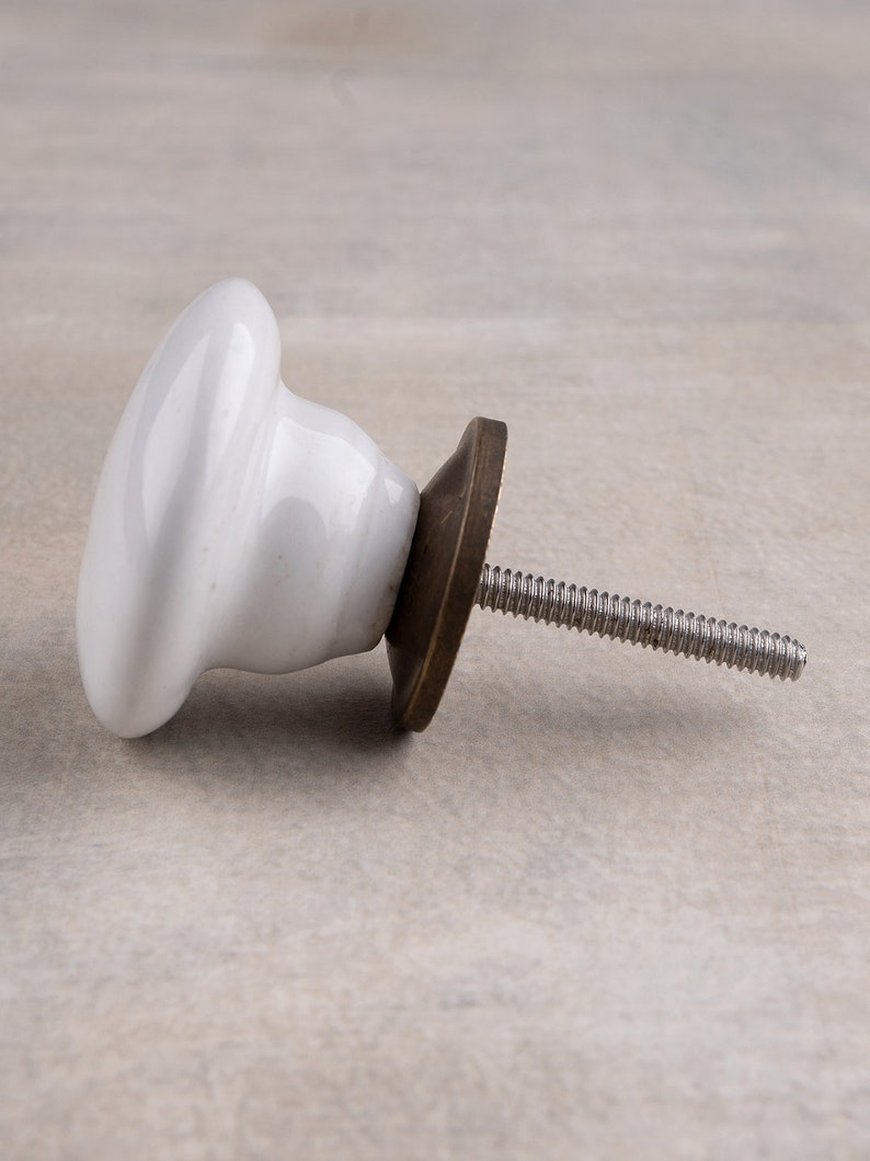 May include: A white ceramic cabinet knob with a smooth, rounded surface and a flared base. The knob features a dark, metallic bronze-toned circular backplate and a long, threaded metal screw extending from the centre for installation purposes.