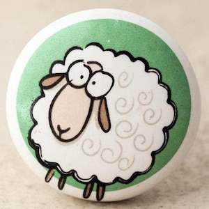 Op de afbeelding: Een witte keramische knop met een groene achtergrond en een cartoon schaap met bruine ogen en poten. Het schaap heeft een wit, pluche lichaam met bruine swirls.