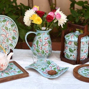 Può includere: Un set di stoviglie in ceramica blu e bianca con motivi floreali. Il set comprende una caraffa, una teiera, un piatto, un piattino e una tazza. Le stoviglie sono decorate con fiori rosa e foglie verdi. Il set è esposto su un tavolo bianco con una base in legno.
