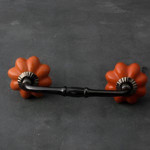 Può includere: Una maniglia per mobili con due pomelli arancioni a forma di zucca e una maniglia nera. I pomelli hanno un design dettagliato e sono attaccati a una barra di metallo nera. L'hardware è progettato per mobili.