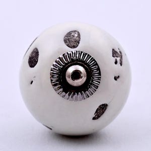 Op de afbeelding: Een ronde, gebroken witte keramische meubelknop met een glanzende afwerking en verspreide, onregelmatige donkergrijze of zwarte vlekken op het oppervlak. Het midden van de knop is voorzien van een cirkelvormig, zilverkleurig metalen beslag.