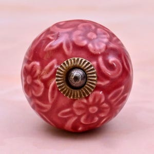 Op de afbeelding: Een ronde, donkerroze keramische meubelknop met een verhoogd, reliëf bloemmotief over het oppervlak. Het midden van de knop is geaccentueerd met een cirkelvormige, messingkleurige metalen plaat met een geribbelde rand en een zilverkleurige schroef.