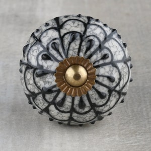 Puede incluir: Un pomo de cerámica blanco con un diseño floral negro y un centro de metal dorado. El pomo tiene una superficie texturizada y un borde elevado.