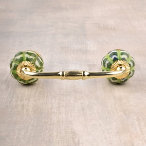 Op de afbeelding: Een gouden metalen kastgreep met twee groene en witte keramische knoppen. De knoppen hebben een geometrisch patroon.