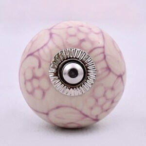 Peut inclure: Un bouton de meuble rond en céramique avec une base de couleur crème ornée d'un motif floral violet clair. Le centre du bouton présente une pièce métallique argentée circulaire avec un bord strié et un trou central pour la fixation.