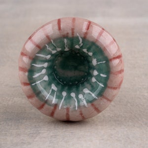 May include: A ceramic knob with a pink and white striped exterior and a green and white interior. The knob has a circular shape and a small hole in the center.