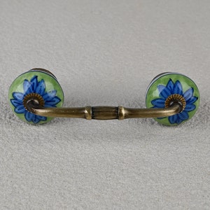May include: A green and blue ceramic drawer pull with a floral design. The pull has a brass bar connecting the two knobs.