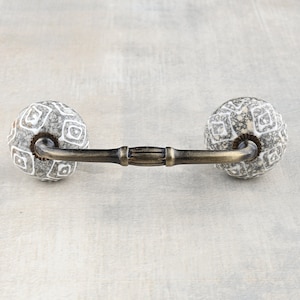 May include: A ceramic cabinet knob with a geometric pattern in white on a gray background. The knob is attached to a brass bar.