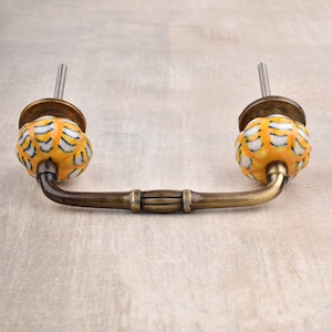 May include: A brass cabinet pull with a yellow and white ceramic knob on each end. The knobs have a floral pattern.