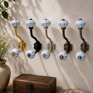 Könnte beinhalten: Fünf Wandhaken mit einem gedrehten Metallarm und zwei weißen Keramikknöpfen mit blauem Blumenmuster. Die Haken sind in Gold, Schwarz, Silber, Antikmessing und Dunkelbronze ausgeführt und verfügen jeweils über eine runde Montageplatte.