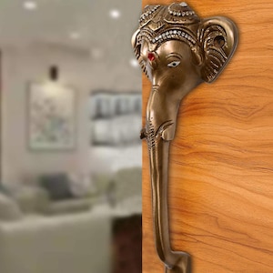 May include: A gold-toned door handle shaped like the head of the Hindu deity Ganesha. The handle has a detailed design with a red gem on the forehead and white eyes.